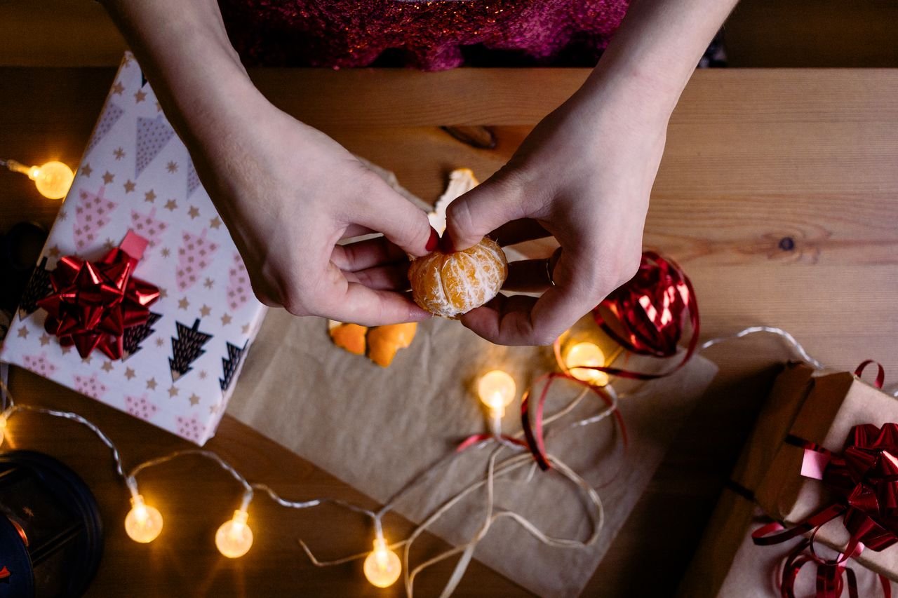 Hands peeling a tangerine amidst Christmas gifts and lights, creating a warm, festive atmosphere.